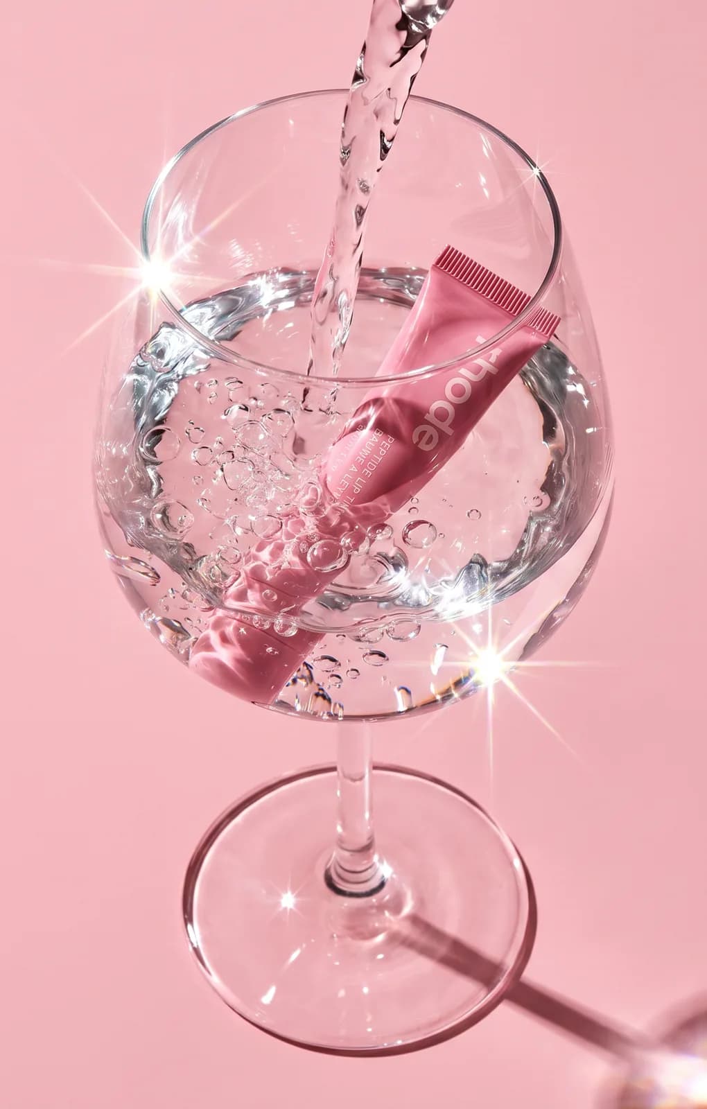 Rhode lip tint submerged in a wine glass as water pours in, pink backdrop